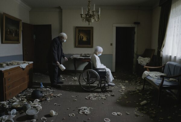 Elderly man and woman in a dimly lit, cluttered room with a wheelchair, evoking isolation.