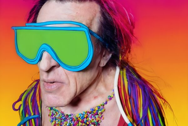 A person in green goggles with colorful hair and a vibrant outfit stands against a vivid pink and orange background.