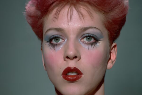 A person with pink hair and bold makeup, featuring blue eyeshadow and red lips, in striking portrait.