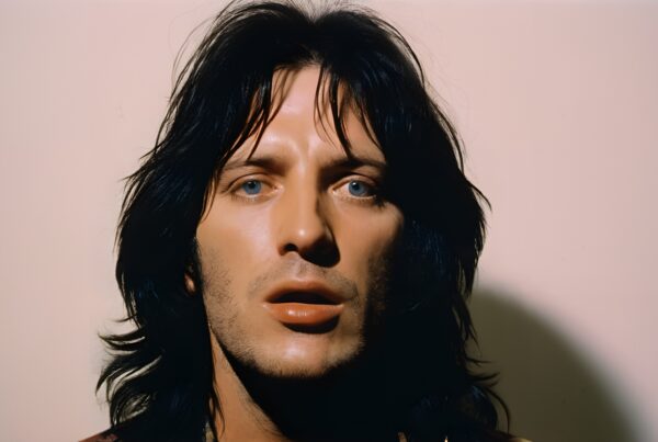 A person with dark hair and blue eyes gazes at the camera against a plain background.