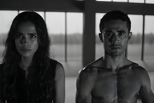 A black and white portrait of a focused man and woman standing indoors, with dramatic lighting and intense expressions.