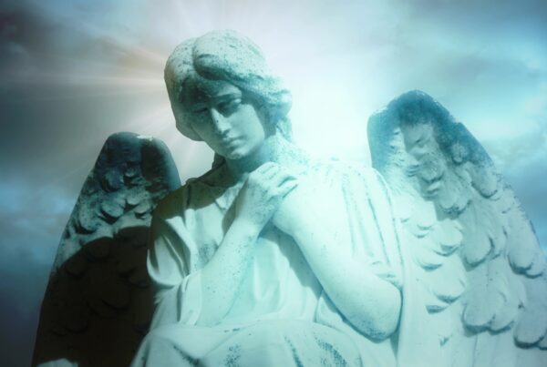 Marble angel statue with serene expression illuminated by radiant light against a backdrop of blue time-lapse clouds and bright sky.