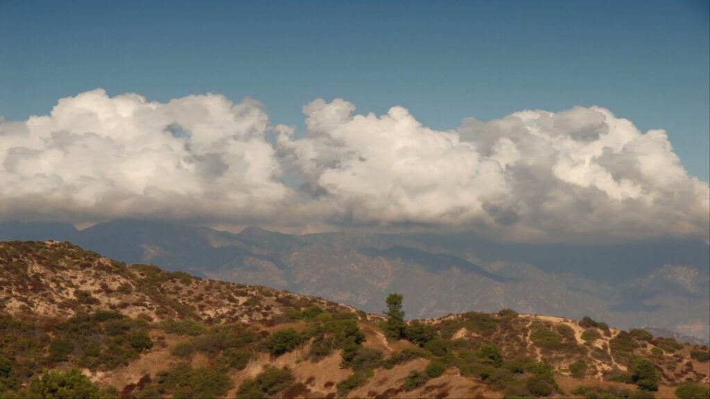 Majestic landscape with fluffy time-lapse clouds over rolling hills and distant mountains under a clear blue sky.