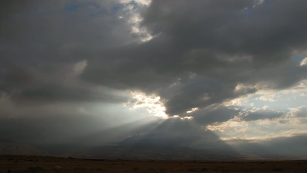 A dramatic time-lapse sky filled with dark clouds, highlighted by sunbeams breaking through, casts light over an arid plain with scattered rocks, set against distant hills.