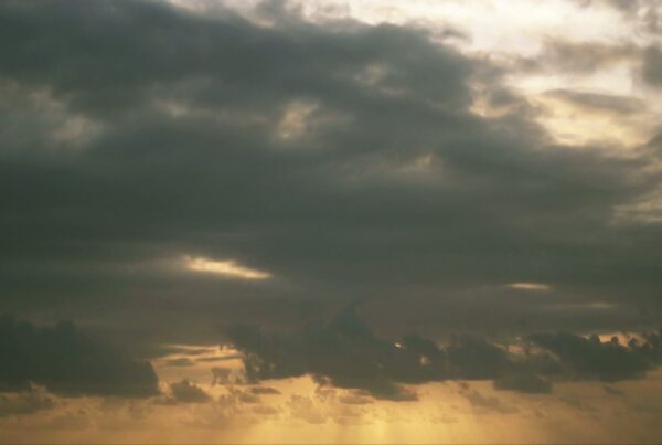Dramatic sky filled with dark time-lapse clouds and beams of sunlight peeking through, casting a golden hue over the horizon, creating a striking contrast and serene yet foreboding atmosphere.