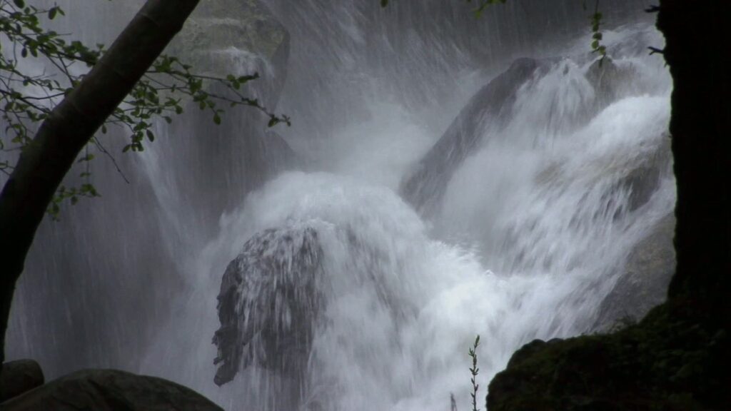 The video captures a powerful waterfall cascading over large rocks. Water is forcefully crashing down, creating a misty aura around the area.