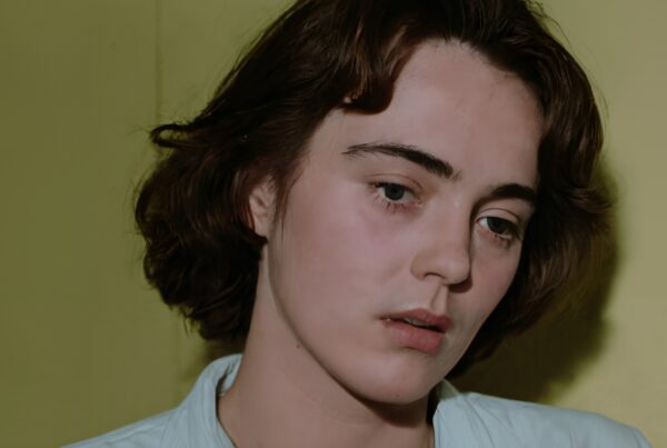 A person with short dark hair looking pensive against a yellow background, wearing a light shirt.