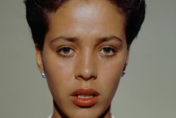 A woman with short dark hair, wearing pearl necklace and earrings, looking directly at the camera with a neutral expression.