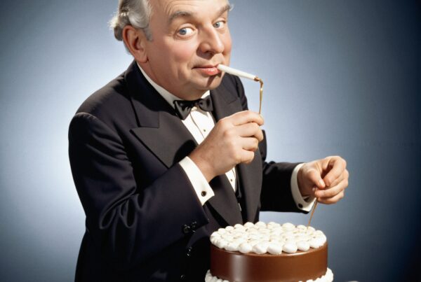 A man in a tuxedo humorously smoking a cigarette above a chocolate cake with white frosting.