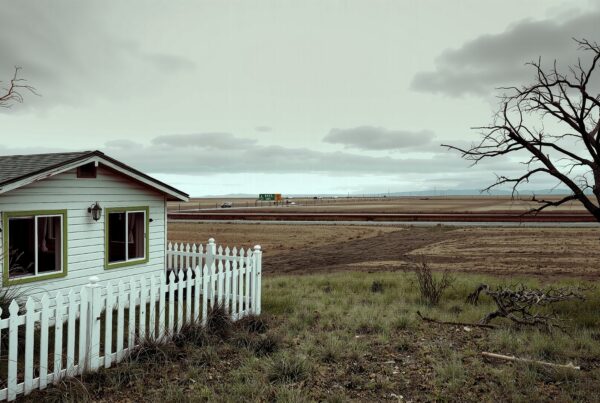 A small white house in a rural landscape beside a highway, with a white picket fence and a leafless tree under cloudy skies.