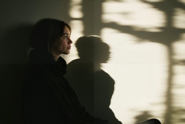 A woman seated in profile, bathed in soft light, with her shadow cast on a wall, creating an artistic and contemplative scene.