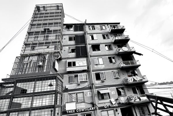 A black and white photo of a multi-story building with open windows, balconies, and a visible sign, framed against a cloudy sky.