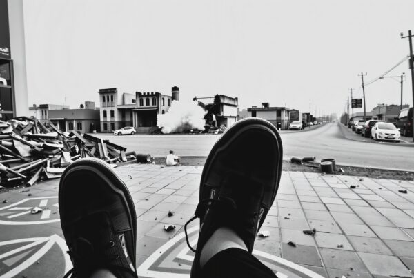 Black and white image showing an urban street scene with debris, smoke, and a person observing from the foreground.