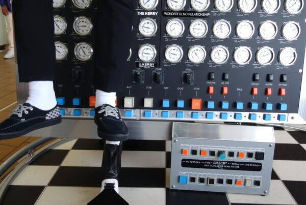 A person sitting on a complex control panel displaying numerous dials and switches in an indoor setting.