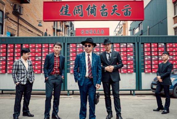 A group of stylish men wearing suits stands confidently against a backdrop of red signs and urban surroundings.