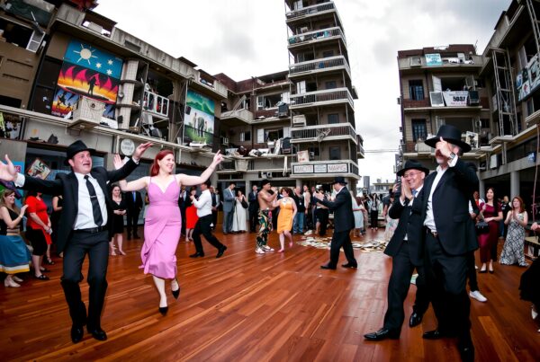 A festive group dances joyfully on a wooden floor in front of a vibrant building facade, under an overcast sky.
