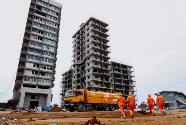 Construction workers on urban site with incomplete leaning buildings, bright orange truck, and scaffolding.