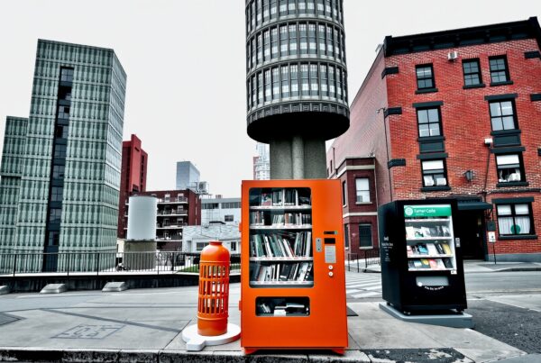 A modern city street features a unique orange book vending machine amidst contrasting architectural elements, blending tradition and innovation.