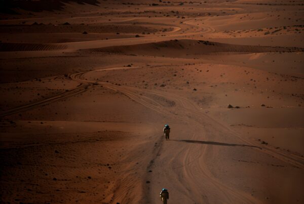 Two motorcyclists journey through a vast desert, casting long shadows on the sand under a clear blue sky.