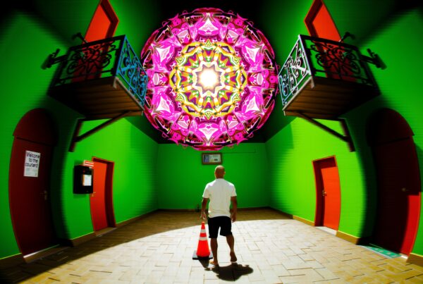 Surreal scene in vivid courtyard with ornate mandala-like structure, colorful walls, and a man holding a traffic cone.