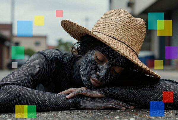 A person in a dotted bodysuit and straw hat resting on an urban street with colorful geometric shapes in the background.