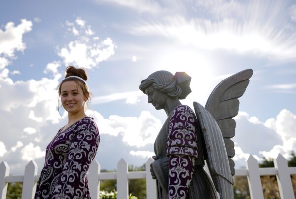 A young woman smiles beside an angel statue in a garden with a blue sky.