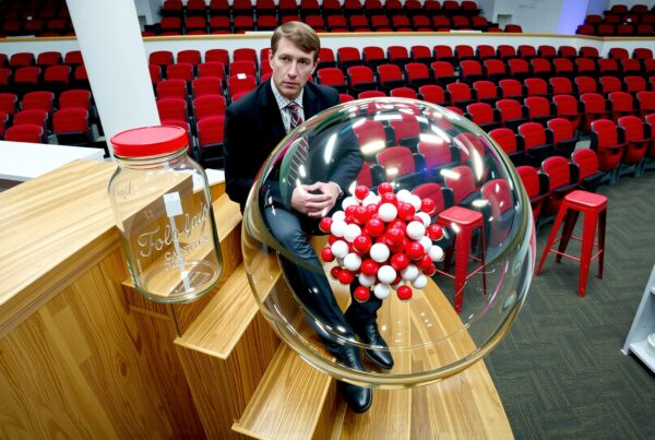 A man in a suit sits near a molecular model and glass jar in a modern auditorium.