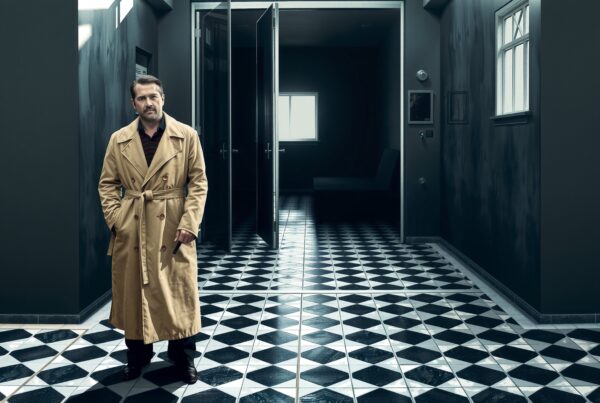 A man in a trench coat stands in a dim hallway with diamond-patterned flooring and partially open doors.