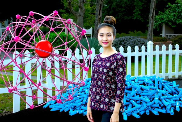 Young woman in patterned shirt stands outdoors with pink geometric sculpture and green garden.