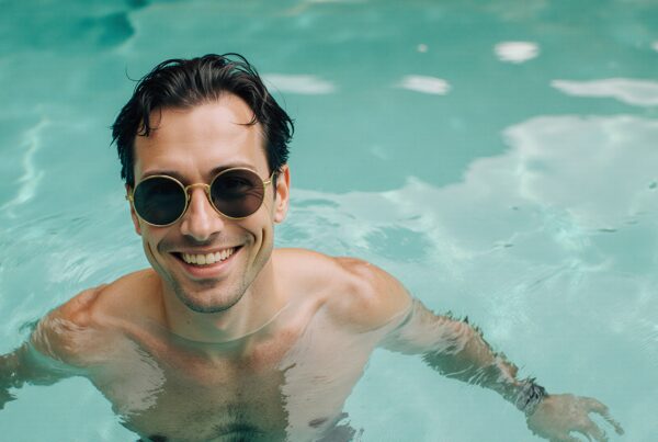 A smiling man in sunglasses enjoys swimming in a serene turquoise pool.