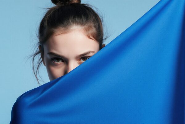 A woman with expressive eyes peers over a vibrant blue fabric in front of a light blue background.