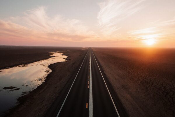 A long highway under a colorful sunset sky with reflections in the water and desert surroundings.