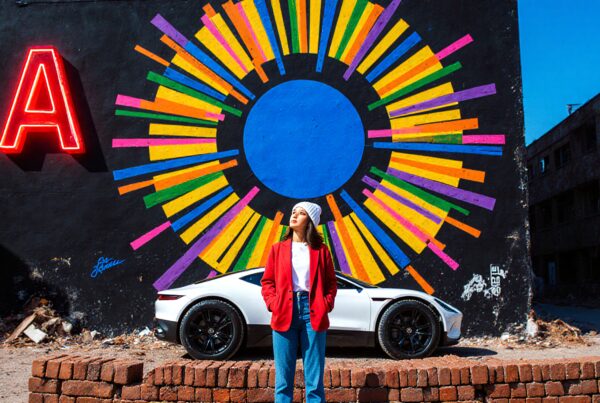 A young woman in a red jacket stands in front of a colorful mural with a white car in an urban setting.