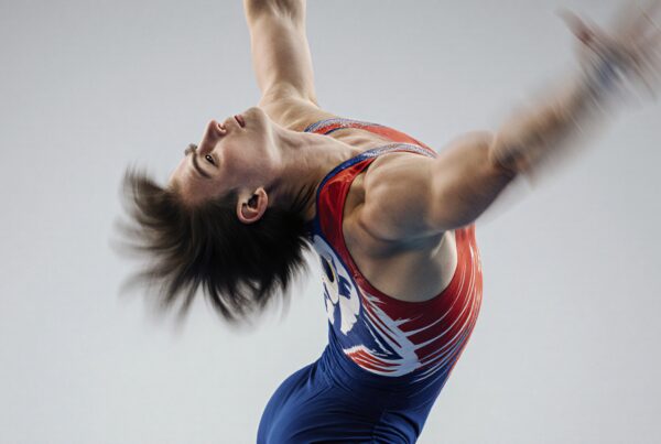 A gymnast in a colorful leotard performs a dynamic leap against a gray background.
