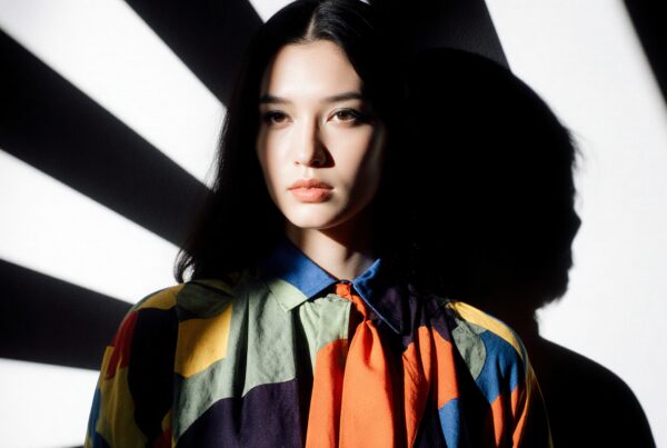 A woman in a colorful blouse stands against a geometric background with dramatic shadows and lighting.