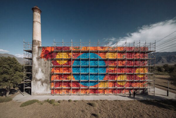 A vibrant mural on a rural industrial structure surrounded by mountains.