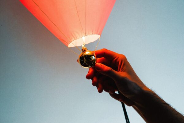 A hand holds a glowing red balloon base with blue sky.