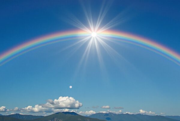 Stunning rainbow arches over lush mountain range beneath vibrant blue sky with glowing sunburst effect