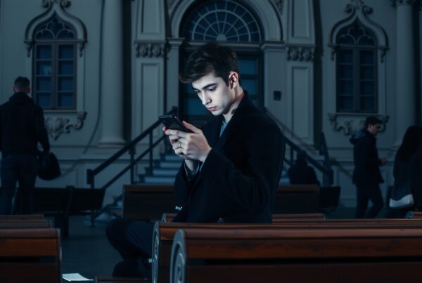 Young man focused on smartphone while sitting on a bench in front of an ornate building in an urban setting.