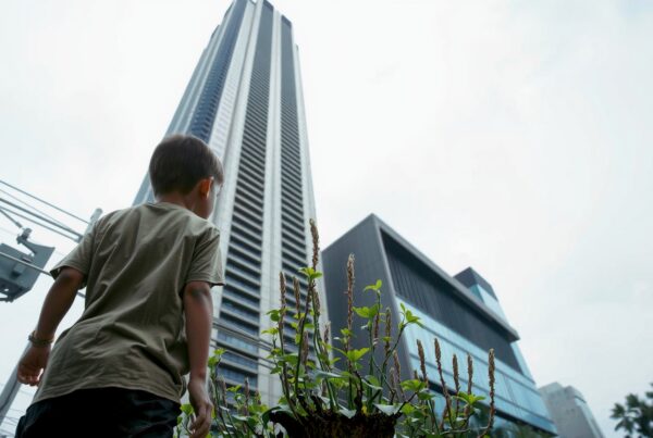 A child looks up at a towering skyscraper amidst green plants in an urban cityscape.