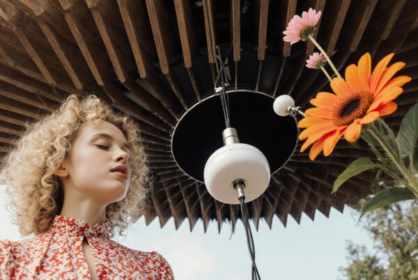 Woman standing under wooden structure, eyes closed in meditation, with vibrant flower in foreground, suggesting harmony between nature and modern design.