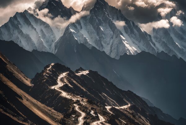 A winding road climbs a rugged landscape to majestic, snow-capped mountains under a dramatic cloudy sky.