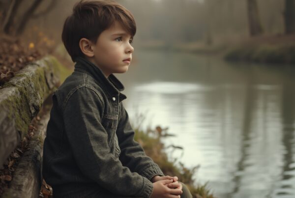 A young boy sits thoughtfully on a rustic bench by a calm river, surrounded by misty trees, in a peaceful autumn setting.