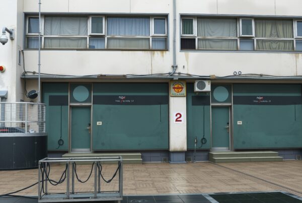 The urban building facade features teal doors, surveillance cameras, and weathered walls.