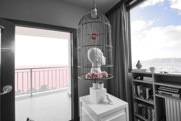 Surreal indoor scene with mannequin head in birdcage, surrounded by roses, near a balcony with mountain view.