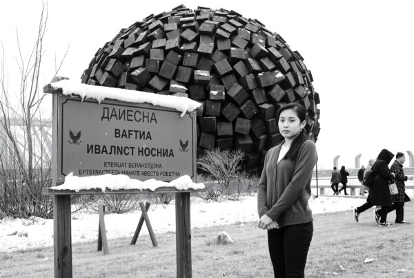 A young woman stands before a geometric sculpture in a snowy, urban park setting with pedestrians passing by.