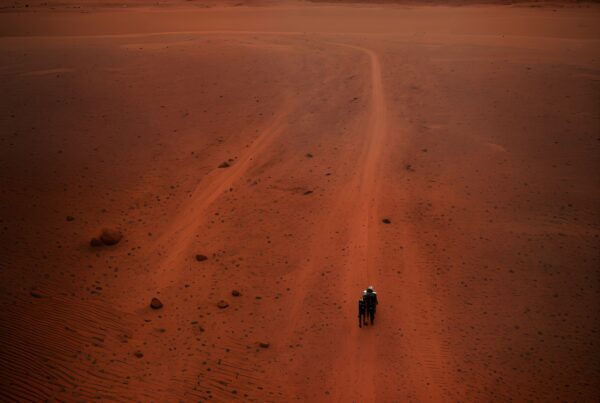 A lone figure walks on a red desert landscape under a setting sun.
