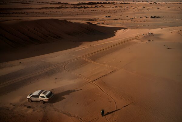 A white SUV and person in a vast desert landscape with dunes and tire tracks.