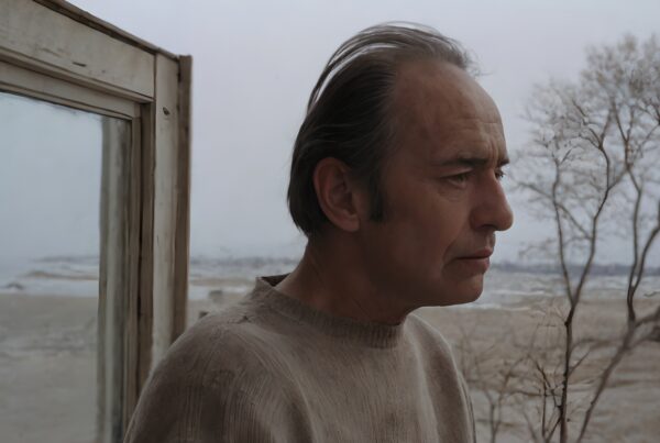 Man in a sweater gazes pensively out a window onto a wintry, barren landscape with frosted trees.
