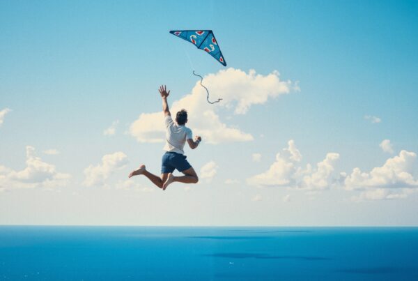 A person jumps while flying a colorful kite against a blue sky and ocean.